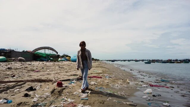 Young woman walking alone on polluted beach with plastic waste wearing eco friendly sustainable clothing sandals and sunglasses, ocean pollution climate change save the planet concept