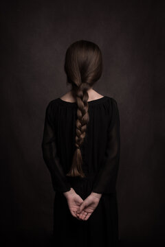 Rear view of girl with long hair in braid and hands on back in classic dark studio portrait