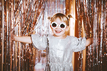 Happy little stylish girl in shiny dress having fun. Festive background with foil curtain...