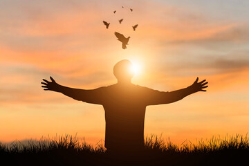 Silhouette of young free man wearing sweater open both arms standing and watched beautiful view sunset alone on top of the mountain with bird flying over the orange sky demonstrate independence.