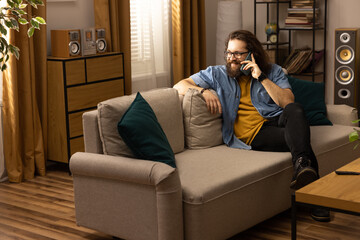 A bearded man in a blue denim shirt is sitting on the living room couch talking on the phone with his girlfriend.