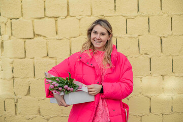Glad shy happy blond woman holding big fresh bouquet of flowers, box gift. Beige brick wall. Congratulations present