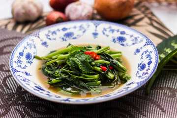 Stir Fried Morning Glory with Soybean Paste Served in a Chinese-patterned dish with spotted leaves, onions, shallots on the table.