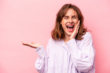 Young caucasian woman isolated on pink background holds copy space on a palm, keep hand over cheek. Amazed and delighted.