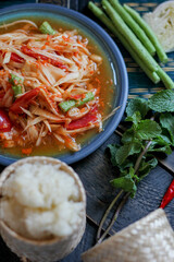 Spicy papaya salad served in a blue dish with sticky rice in a bucket and long beans, dried chilli and vegetables.