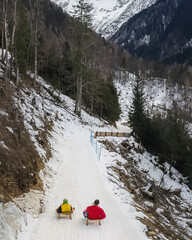 Man and child sledding side by side, on a narrow mountain snowy road along the steep slope, aerial view.