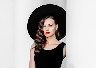 High fashion outdoor full length portrait of elegant woman in black hat and dress standing behind white pillar. Vintage-style portrait of a woman in black with red lips. - obrazy, fototapety, plakaty