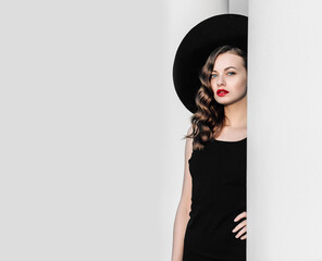 High fashion outdoor full length portrait of elegant woman in black hat and dress standing behind white pillar. Vintage-style portrait of a woman in black with red lips.