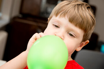 3 year old boy inflating green balloon with a mouth. Swollen cheeks and bulging eyes. Lungs, breathing exercise. Pasty accessories.