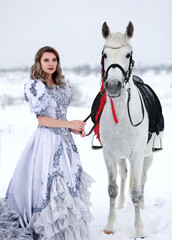 Fototapeta na wymiar Young woman in light blue ball gown with white horse in winter field. Winter fairytale. Fantasy portrait of a girl