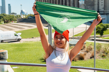 Beautiful young woman activist raising a green scarf that symbolizes the feminist fight for...