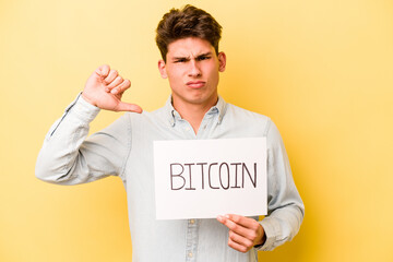 Young caucasian man holding bitcoin placard isolated on yellow background showing a dislike...