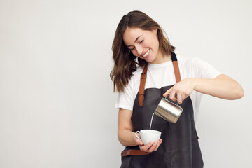Professional smiling barista woman in studio serving coffee, pour milk from a jug in a coffee cup. Café latte. Isolated on white background. 