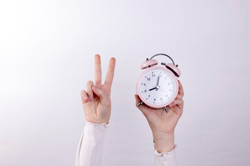Human arm raised up holding pink alarm clock and showing victory symbol on white background....