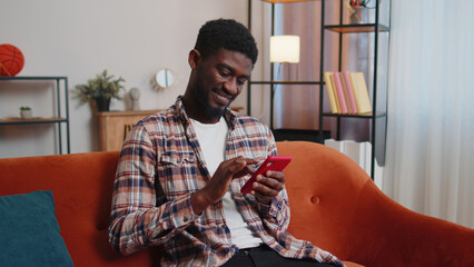 Joyful African American young man in casual shirt sitting on sofa, using smartphone or mobile cell phone in modern room at home. Tapping, scrolling, watching video, blogger content, online shopping
