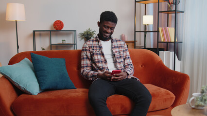 Cheerful relaxing young african american man sitting on sofa uses mobile phone smile at modern home apartment. Happy guy texting share messages content on smartphone social media applications online