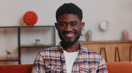Obraz premium Portrait of happy one african american teenager student guy in casual shirt smiling, looking at camera. Young cute fashion model man indoors isolated at home in living room on sofa. Male nature beauty