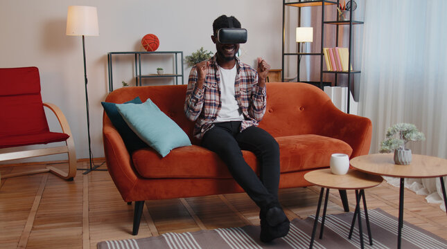 African American man wearing VR headset resting at home sitting on sofa in living room alone using futuristic technology making gestures with hands in air, play 3D video game. Virtual reality glasses