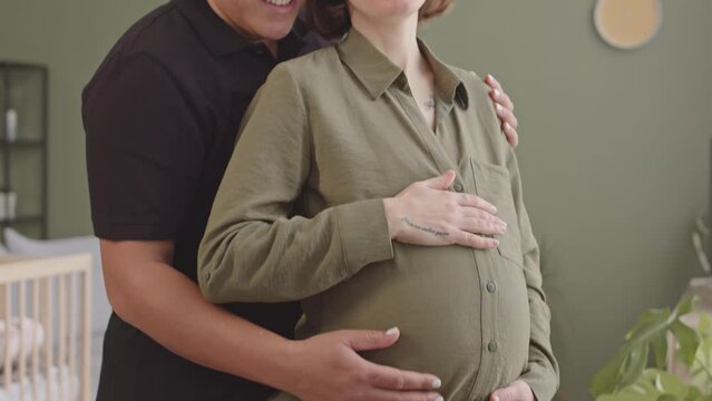 Tilt slowmo of short haired Biracial woman stroking belly of her pregnant wife, standing together in embrace at living room and smiling