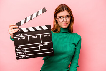 Young caucasian woman holding a clapperboard isolated on pink background confused, feels doubtful...