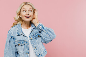 Elderly curious nosy happy woman 50s wearing denim jacket try to hear you overhear listening intently isolated on plain pastel light pink background studio portrait. People lifestyle fashion concept.