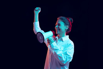 Half-length portrait of young pretty girl in white shirt shouting at megaphone isolated on dark background in blue neon light, filter. Concept of emotions, fashion, youth