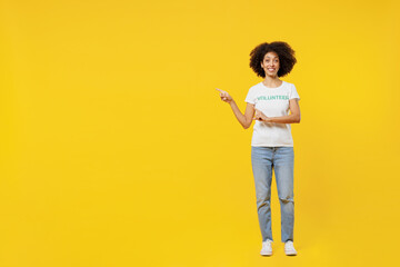 Full body young woman of African American ethnicity in white volunteer t-shirt point index finger aside workspace area isolated on plain yellow background. Voluntary free work assistance help concept.