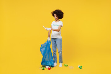 Full length young woman of African American ethnicity wears white volunteer t-shirt hold trash bag clean remove rubbish isolated on plain yellow background Voluntary free work assistance help concept