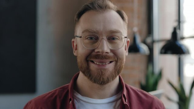 Portrait of the skillful bearded male programer looking at the camera in cafe with pleasure smile. Adult freelancer in casual clothes posing on camera while sitting at the table