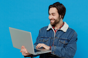 Smiling happy cheery young brunet bearded man 20s wear warm denim jacket hold use work on laptop pc computer isolated on plain pastel light blue background studio portrait. Winter cold season concept