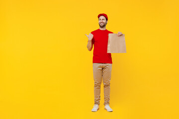 Full body delivery guy employee man in red cap T-shirt uniform workwear work as dealer courier hold brown blank craft paper takeaway bag mock up point finger aside isolated on plain yellow background.