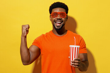 Young smiling fashionable fun happy man of African American ethnicity 20s wear orange t-shirt stylish red glasses hold cup of soda pop fizzy water isolated on plain yellow background studio portrait