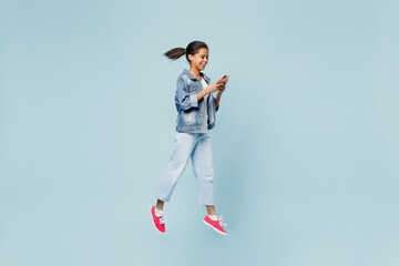 Full body fun little kid teen girl of African American ethnicity 12-13 years old in denim jacket jump high hold use mobile cell phone isolated on pastel plain light blue background. Childhood concept.
