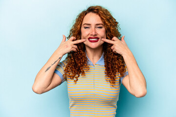 Fototapeta premium Young ginger caucasian woman isolated on blue background smiles, pointing fingers at mouth.