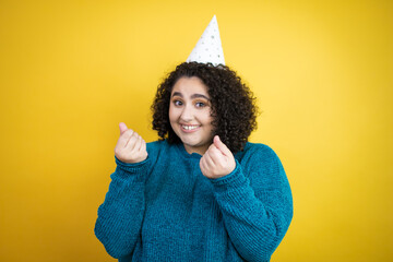 Young beautiful woman wearing a birthday hat over isolated yellow background doing money gesture...
