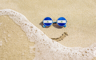 Fototapeta na wymiar Sunglasses with flag of Nicaragua on a sandy beach. Nearby is a sea lightning and a painted smile. The concept of a successful vacation in the resorts of Nicaragua.