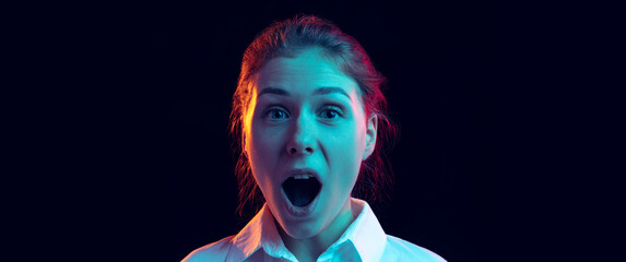 Shocked young girl in white shirt looking at camera with open mouth isolated on dark background in...