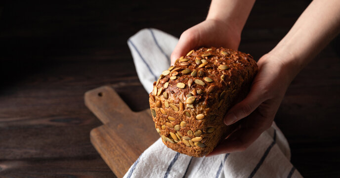 Homemade crusty loave of bread with pumpkin seeds on wooden background. Dark mood. Baker holding fresh bread in the hands. Traditional techniques, innovating bread, slow carb baking