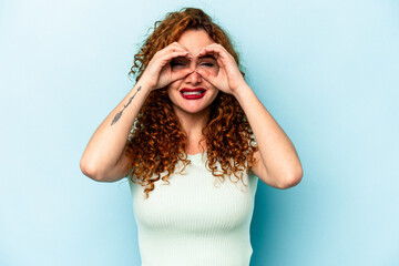 Young ginger caucasian woman isolated on blue background showing okay sign over eyes