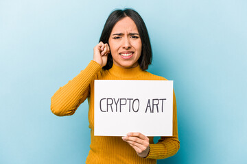 Young hispanic woman holding crypto art placard isolated on blue background covering ears with hands.