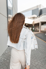 Fashion young brunette woman with stylish blue denim jacket walks on the street, back view