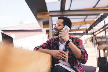 Cheerful male in optical glasses for provide eyes correction enjoying mobile communication for talking, happy Hispanic hipster guy with takeaway coffee to go using cellphone for making conversation