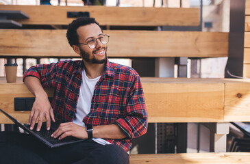 Cheerful millennial hipster guy in spectacles enjoying freelance job using netbook for online...