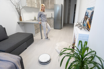 young woman using automatic vacuum cleaner to clean the floor, controlling smart machine housework...