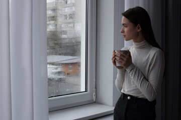 Fototapeta na wymiar Melancholic young woman with drink looking out of window on rainy day, space for text. Loneliness concept