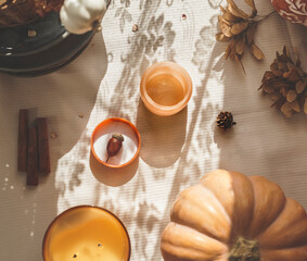 Cozy still life with candles, pumpkin, cinnamon sticks and autumn leaves at beige table blanket with sunlight. Seasonal hygge decoration with shadow. Top view.