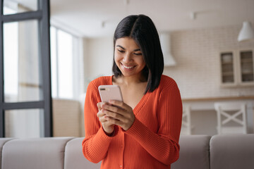 Young beautiful African American woman wearing casual clothing holding smartphone text messaging, communication online sitting at home. Smiling hipster female using mobile app shopping online