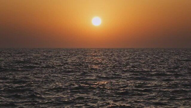 Orange sky and sun setting over calm waves of a gray sea or ocean. Tranquil, zen-like, cinematic scene. Slow motion video.