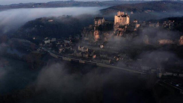 Beautiful cinematic aerial view of Beynac castel above the fog and river in sunrise light, Dordogne France, blue mood