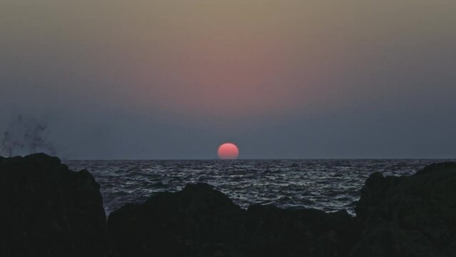 Big red sun setting slowly over the sea or ocean horizon, grey sky with slow motion waves, rocky coast. Calm, zen-like scene. Montenegro landscape.